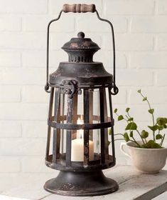 an old metal lantern with two lit candles in it on a table next to a potted plant