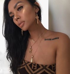 a woman with long black hair wearing gold necklaces and matching earring, standing in front of a white wall