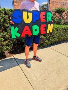 a man holding a sign that says super kaden in front of a building with bushes