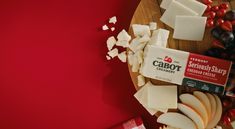 cheese and fruit on a wooden cutting board next to a red tablecloth with the word cabot printed on it