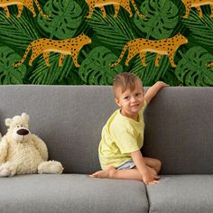 a little boy sitting on a couch next to a teddy bear and wallpaper with giraffes