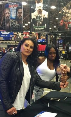 two women are shaking hands in front of a table at an indoor game show with posters on the wall behind them