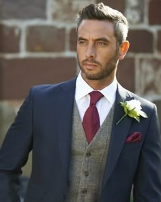 a man wearing a suit and tie with a flower in his lapel, standing next to a brick wall