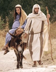 a man and woman dressed in biblical garb walking down a dirt road with a donkey