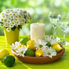 a candle and some lemons in a bowl on a yellow table cloth with daisies