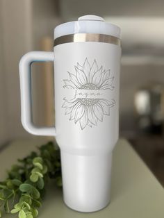 a white coffee cup sitting on top of a table next to green leaves and a plant