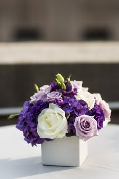 purple and white flowers sit in a square vase on a round table with an elegant centerpiece