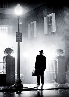 a man in a hat and coat is standing on the street at night with fog