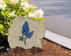 a rock with a blue flower painted on it sitting in the dirt near some flowers