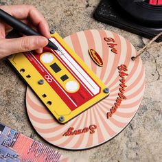 a hand holding a pen over a yellow and red tape cassette shaped coaster with the words'80s'written on it