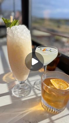 two glasses filled with drinks sitting on top of a table next to each other near a window