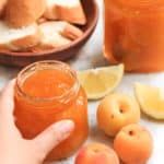 a person is holding a jar of peach jam with lemons and bread in the background