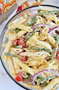 a pasta salad with asparagus and tomatoes in a glass bowl on a marble table