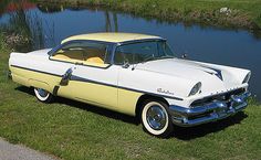 an old yellow and white car parked next to a body of water in the grass