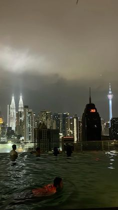 people are swimming in the water at night with city lights and skyscrapers behind them