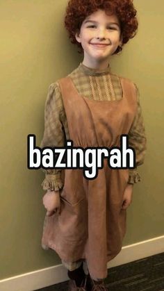 a young boy wearing a brown dress standing next to a wall with the words bazinggrah on it