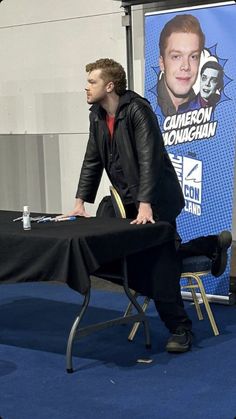 a man sitting at a table in front of a blue backdrop with a poster on it