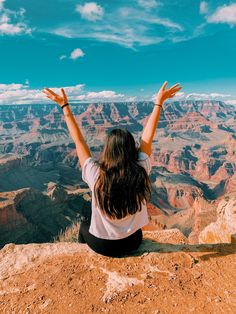 a woman sitting at the edge of a cliff with her hands up in the air