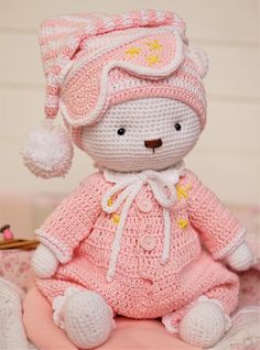 a crocheted teddy bear wearing a pink dress and hat sitting on a bed