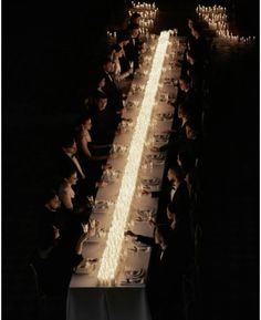 a long table with many people sitting at it and lit candles in the dark behind them