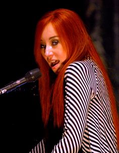 a woman with long red hair and striped shirt playing the piano in front of a microphone