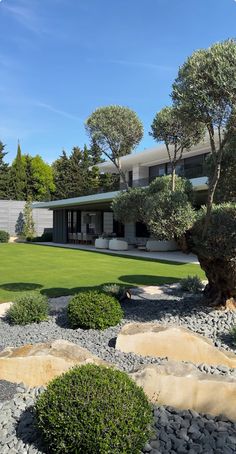 an outdoor area with grass, rocks and trees in front of a modern house on a sunny day