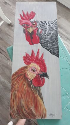 a painting of roosters is being held up by someone's hand on a table