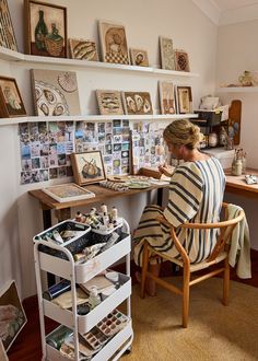 a woman sitting at a desk with lots of pictures on the wall behind her and she is painting