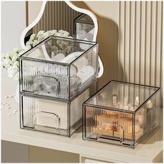 two clear storage containers on top of a white table next to a mirror and flowers