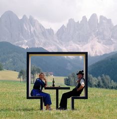 two people sitting at a table in the middle of a field with mountains behind them