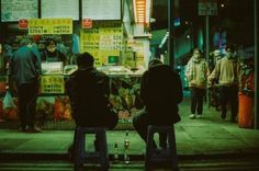 two people sitting on stools in front of a food stand at night with neon lights