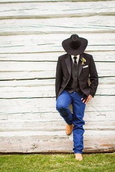 a man in a suit and hat is leaning against a wooden wall with his hands on his hips