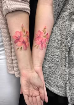 two women holding hands with matching tattoos on their arms and both have pink flowers on them