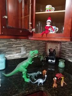 an elf is sitting in the kitchen with toy dinosaurs on the counter and shelves above