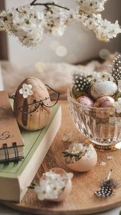 an arrangement of eggs and flowers in a glass bowl on a wooden tray next to two books