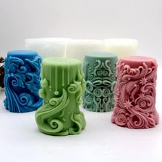 four different colored candles sitting next to each other on a white surface with pine cones in the background