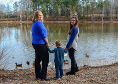 two women and a boy are standing by the water holding hands with ducks in the background