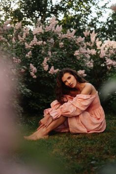 a woman sitting in the grass with her legs crossed and wearing a pink dress next to flowers