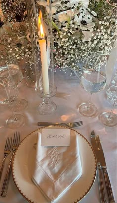 a table set with silverware and white napkins, candles and flowers in the background