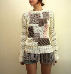 a woman standing in front of a white wall wearing a sweater with fringes on it