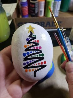 someone is holding an easter egg with a christmas tree painted on it and a paintbrush in their hand