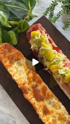 two long sandwiches sitting on top of a wooden cutting board next to green plant life