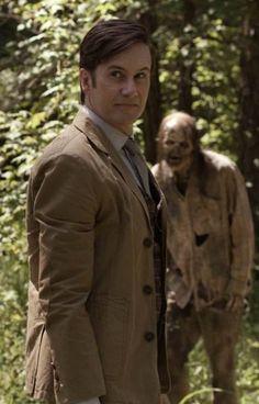 a man in a suit standing next to a dead animal on the forest floor with another person behind him