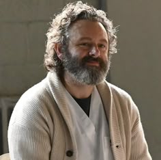 a man with long hair and beard wearing a cardigan sitting in front of a window