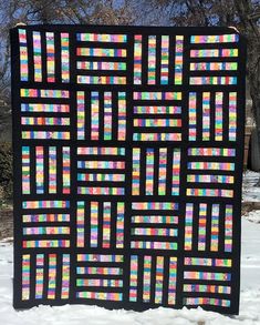 a black quilt with multicolored squares on it in the snow next to some trees