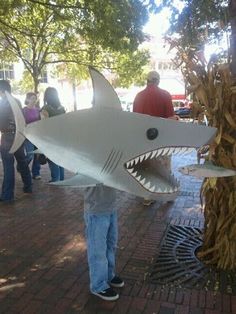 a man holding up a fake shark head in front of a tree with people standing around it