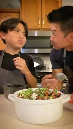 a man standing next to a little boy in front of a bowl of food on top of a counter