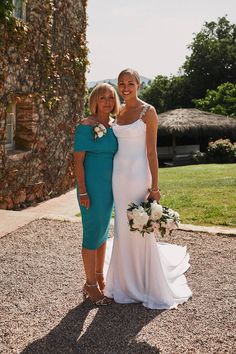 two women standing next to each other in front of a stone wall and grass field