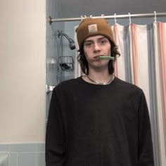 a man standing in front of a shower with a toothbrush in his mouth and wearing a hat