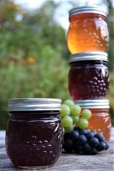 three mason jars filled with different types of jam and grapes sitting on a wooden table